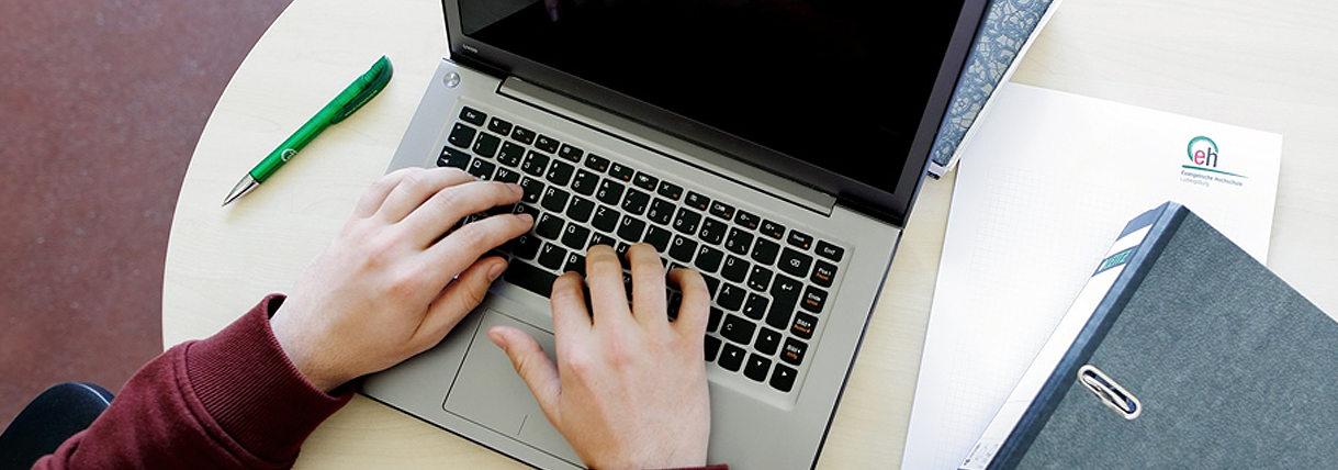 Blick von oben auf einen Arbeitsplatz zeigt Hände auf einer Laptop-Tastatur beim Tippen. Neben dem Laptop auf dem Schreibtisch ist ein Ordner, sowie ein Notizblock mit E.H. Ludwigsburg Logo zu sehen.